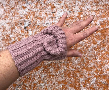 Hand mit altrosa gehäkelter, fingerloser Armstulpe mit geripptem Muster und rundem Zierakzent