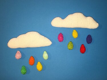 Zwei weiße gehäkelte Wolken mit bunten gehäkelten Regentropfen vor blauem Hintergrund