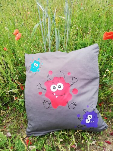 Graues Kissen mit drei bunten Klecks‑Monstern in Rot, Lila und Blau auf einer Wiese