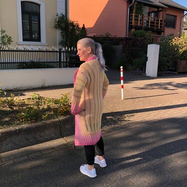 Rückansicht: Person trägt langen, gehäkelten Seelenwärmer in Gelb und Pink über schwarzen Hosen und weißen Schuhen auf einem Gehweg