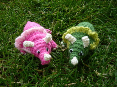 Zwei gehäkelte Triceratops-Amigurumi, einer pink, einer grün, auf Gras
