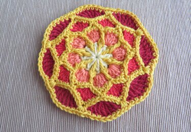 Runder gehäkelter Untersetzer mit gelbem Gitter, rosa und korallfarbenen Segmenten und weißem Zentrum auf hellem Stoff