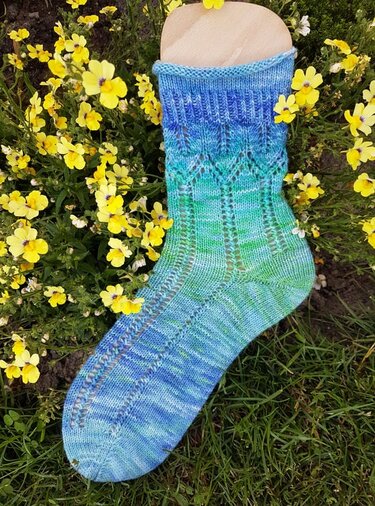 Blau-grün gestrickte Damensocke mit Ajourmuster auf einem Holzsockenformer zwischen gelben Blumen