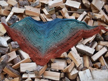 Gehäkeltes Dreieckstuch mit Farbverlauf von Blaugrün bis Rost und Spitzenmuster auf Holzstapel