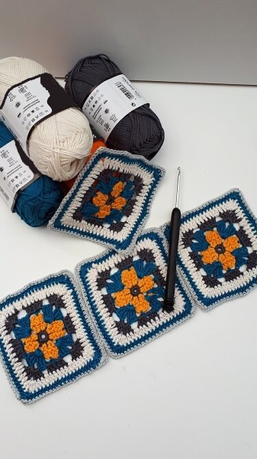 Mehrere gehäkelte Granny-Squares mit senfgelber Blumenmitte, petrol-, dunkelgrauem und weißem Rahmen, Garnknäueln und Häkelnadel