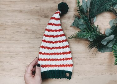 Gestreifte Zipfelmütze in Rot, Weiß und Grün mit Bommel, auf Holzhintergrund von einer Hand gehalten, rechts ein grüner Kranz