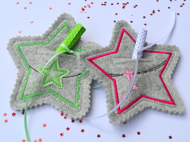 Zwei graue Filzsterne mit grüner und pinker Stickkante, jeweils mit kleinem Fach, Bändern und Sternkonfetti