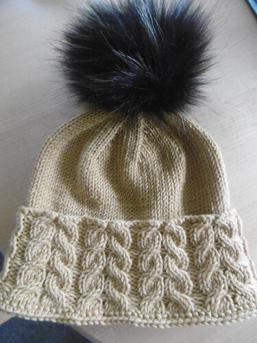 Beigefarbene Strickmütze mit Zopfmuster, glatt rechts gestricktem Körper und großem schwarzem Bommel.