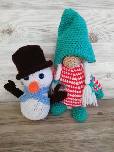 Zwei gehäkelte Amigurumi: Schneemann mit braunem Zylinder und blauem Schal sowie ein Wichtel mit türkisfarbener spitzer Mütze und rot-weiß gestreifter Kleidung auf hellem Holzuntergrund.