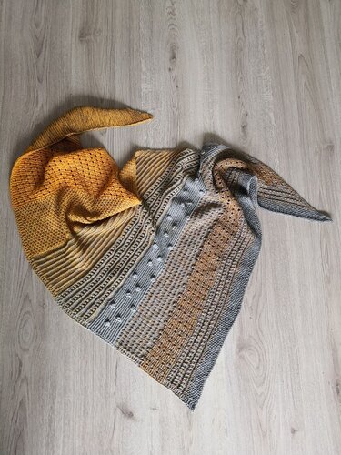 Gestricktes Dreieckstuch in Gelb- und Grautönen mit Streifen- und Noppenmustern auf hellem Holzfußboden