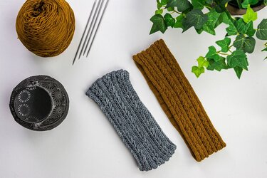 Zwei gestrickte Stirnbänder in Grau und Rostbraun mit Wollknäuel, Stricknadeln und einer Pflanze auf hellem Hintergrund.