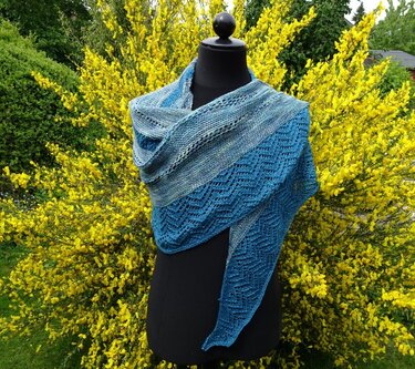 Blaues gestricktes Halbrundtuch mit Wellen-Ajour auf schwarzer Schneiderpuppe vor gelbem Blühstrauch