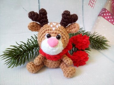 Small brown crochet reindeer amigurumi with red pom-pom scarf and pine sprig