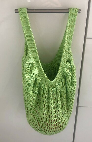 Hellgrüne gehäkelte Netztasche mit breiten Henkeln an einer Stange vor hellem Hintergrund.