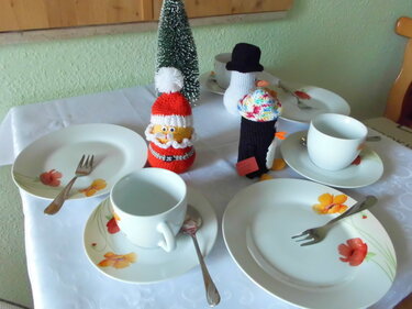 Tisch mit drei gehäkelten Weihnachtsfiguren (Weihnachtsmann, Pinguin, Schneemann), Blumen-Geschirr und kleinem Weihnachtsbaum.