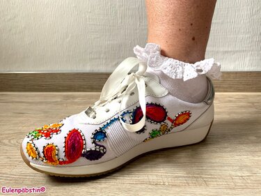 Nahaufnahme eines weißen Sneakers mit bunten textilen Verzierungen und einer weißen Socke mit gehäkeltem Rüschenabschluss.