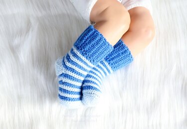 Pair of blue and white striped crochet baby socks showing ribbed cuff and heel shaping on a white fuzzy surface