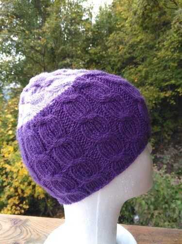 Gestrickte Mütze in Lila und Hellviolett mit Strukturmuster auf weißem Schaufensterkopf vor verschwommenem Grün.