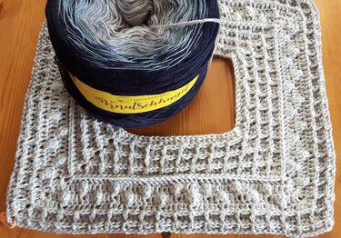 Farbverlaufsgarn in Blau- und Grautönen neben einem grauen, gehäkelten Stück mit quadratischer Aussparung und strukturiertem Muster.