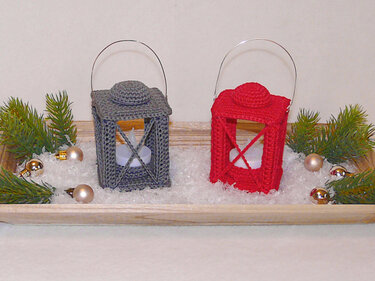 Two small crocheted lanterns in red and gray on a wooden tray with faux snow, pine sprigs, baubles and tea lights
