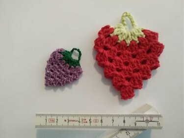 Zwei kleine gehäkelte Früchte (Erdbeere und Traube) auf hellem Hintergrund, mit Lineal zur Größenreferenz.