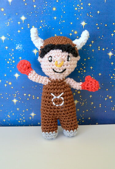Kleines gehäkeltes Amigurumi-Stier mit Hörnern und Stierzeichen, braune Latzhose und rote Handschuhe vor blauem Sternenhintergrund