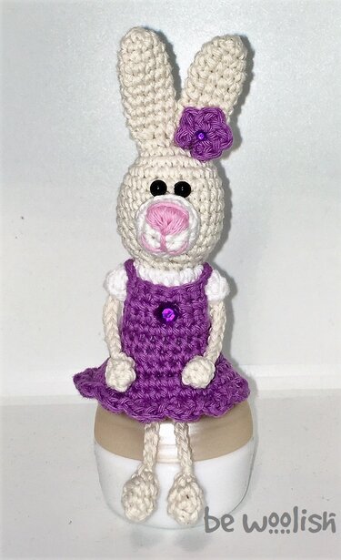 Gehäkelter Amigurumi-Hase in cremeweiß mit lila Kleid und Blume, sitzend