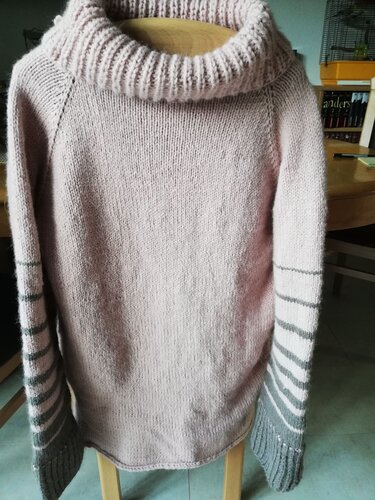 Gestrickter rosa Raglanpullover mit hohem Rollkragen und grau-rosa gestreiften Ärmelbündchen, drapiert auf einer Stuhllehne.