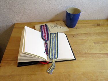 Zwei gehäkelte Lesezeichen mit Quasten in einem aufgeschlagenen Buch auf einem Holztisch