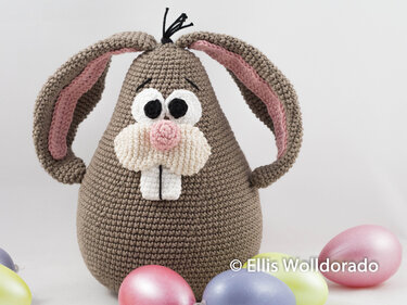 Gehäkelter brauner Hase mit rosa Innenohren und weißen Zähnen neben bunten Ostereiern auf hellem Hintergrund.
