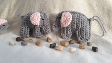 Zwei graue Elefanten-Amigurumi mit rosa Ohren und einer Garnschlaufe auf hellem Hintergrund, davor glatte Steine.