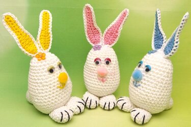 Drei weiße Amigurumi-Osterhasen mit gelben, rosa und blauen Innenohren, Wackelaugen und Pompon-Nasen vor hellgrünem Hintergrund.
