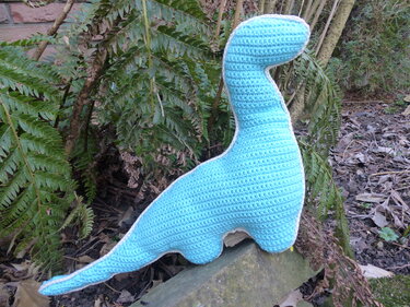 Hellblauer gehäkelter Amigurumi-Dinosaurier mit weißer Umrandung auf einem Stein im Freien
