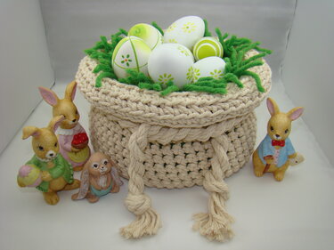 Beigefarbener gehäkelter Korb mit Kordelzug, gefüllt mit weiß-grünen Ostereiern auf grünem Garn, umgeben von Hasenfiguren