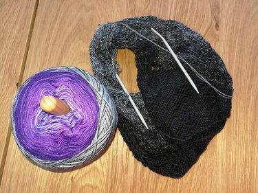 Lila-graues Verlaufsgarn mit Holzkern und ein begonnenes Dreieckstuch mit Rundstricknadeln auf einer Holzoberfläche.