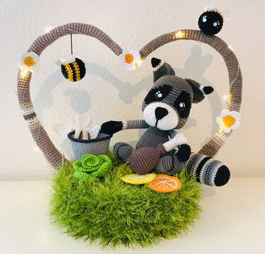 Gehäkelter Waschbär-Amigurumi sitzt in einem Herzrahmen mit Blüten, Lichterkette, hängender Biene und gehäkelten Details auf grünem Untergrund