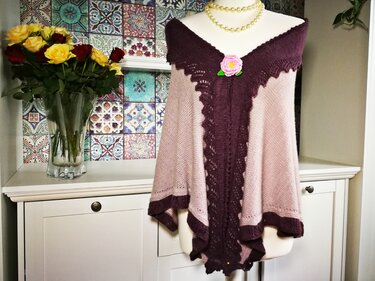 Gestricktes Rechtecktuch in Rosé und Dunkellila mit mittigem Lochmuster, gewellten Kanten und gehäkelter Blumenbrosche auf einer Schneiderpuppe.