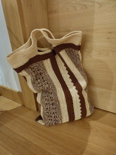 Beige-braune gehäkelte Tasche mit Streifen und Henkeln lehnt an einer Holztür auf Holzboden