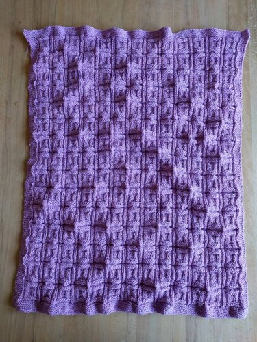 Pastelllila gestrickte Babydecke mit Relief-Karomuster und kraus gestricktem Rand auf Holzboden