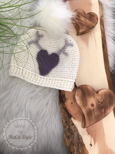 Cremefarbene gehäkelte Babymütze mit lila Herzapplikation, hellgrauen Geweih-Stichen und weißem Kunstfell-Bommel neben Holzherzen
