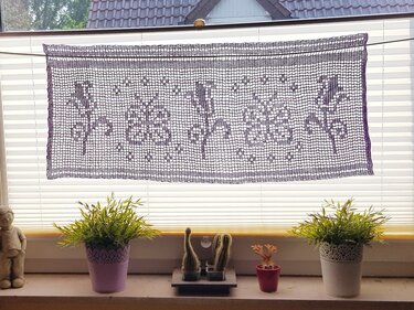 Dunkelviolette gehäkelte Filetgardine mit Blumen- und Rankenmuster vor einem Fenster, darunter Pflanzen auf der Fensterbank.