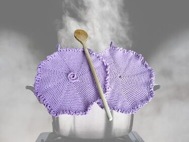 Zwei lila gehäkelte Topflappen und ein Holzlöffel auf einem dampfenden Kochtopf