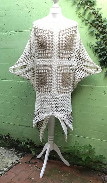 Gehäkeltes Granny‑Square‑Kleid in Creme und Hellbraun auf einer Schneiderpuppe vor grüner Wand mit Efeu; offene Maschen und weiter Schnitt sichtbar.