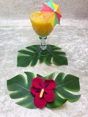 Gehäkelte rote Hibiskusblüte mit gelbem Stempel und violetten Akzenten auf grünen, tropischen Blättern; im Hintergrund ein Glas mit orangefarbenem Getränk und buntem Papier-Sonnenschirm.
