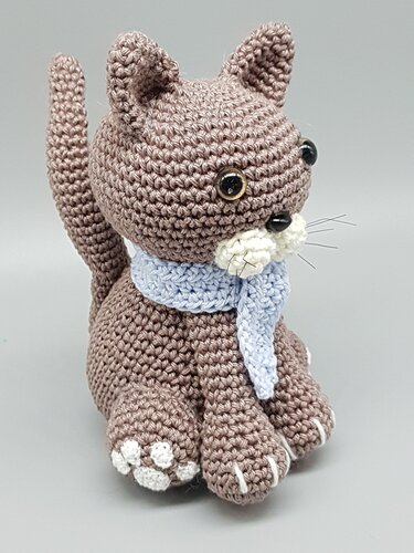 Braune gehäkelte Amigurumi-Katze mit hellblauem Schal und weißen Pfoten vor grauem Hintergrund