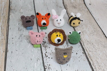 Acht kleine gehäkelte Tierköpfe: Bär, Fuchs, Hase, Biene, Schwein, Löwe, Frosch und Eule auf hellem Holzuntergrund.