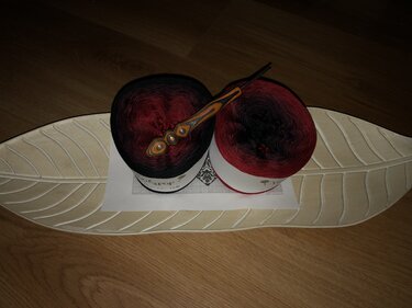 Zwei Bobbel aus rot‑schwarzem Verlaufsgarn und eine verzierte Häkelnadel auf einer Häkelanleitung und einem blattförmigen Tablett