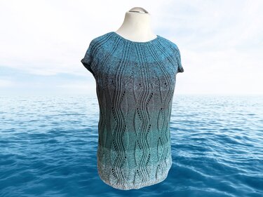 Gestricktes Damenshirt mit kurzen Ärmeln, blau-türkis-grauem Farbverlauf und Wellen-Lochmuster auf Schneiderpuppe vor Meereshintergrund.