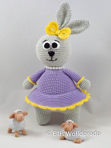 Gehäkelter grauer Amigurumi-Hase im lila Kleid mit gelber Schleife und zwei kleinen Schafen