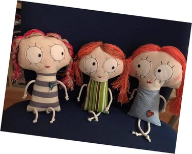 Drei handgemachte Stoffpuppen mit roten Garnhaaren, gestickten Augen und unterschiedlichen Kleidern sitzen nebeneinander auf dunkelblauem Untergrund.